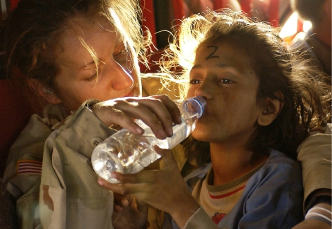 A child getting help from a female army officer to drink water in a disaster scene.